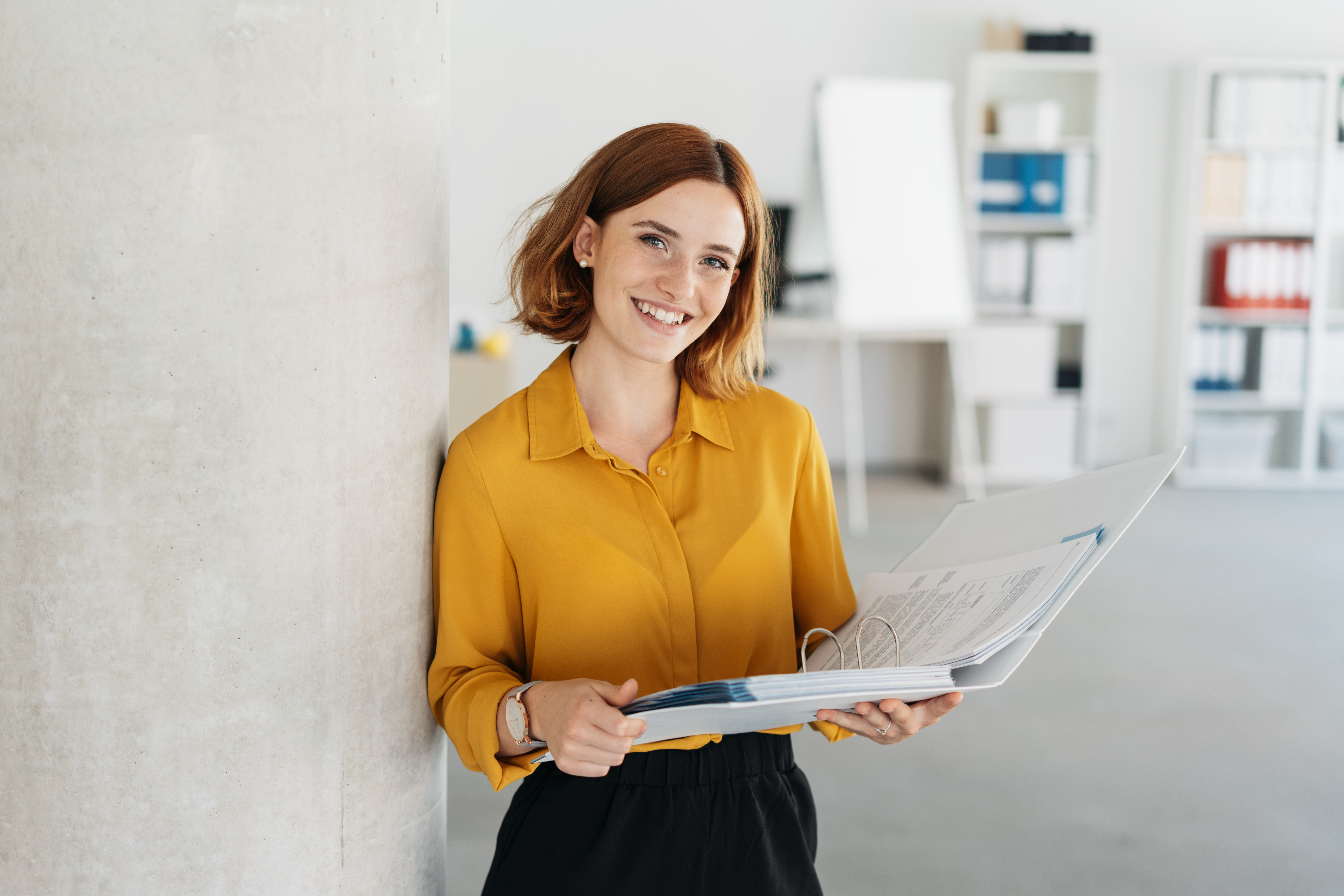 smiling-woman-in-yellow-blouse-holding-binder-in-office.jpeg