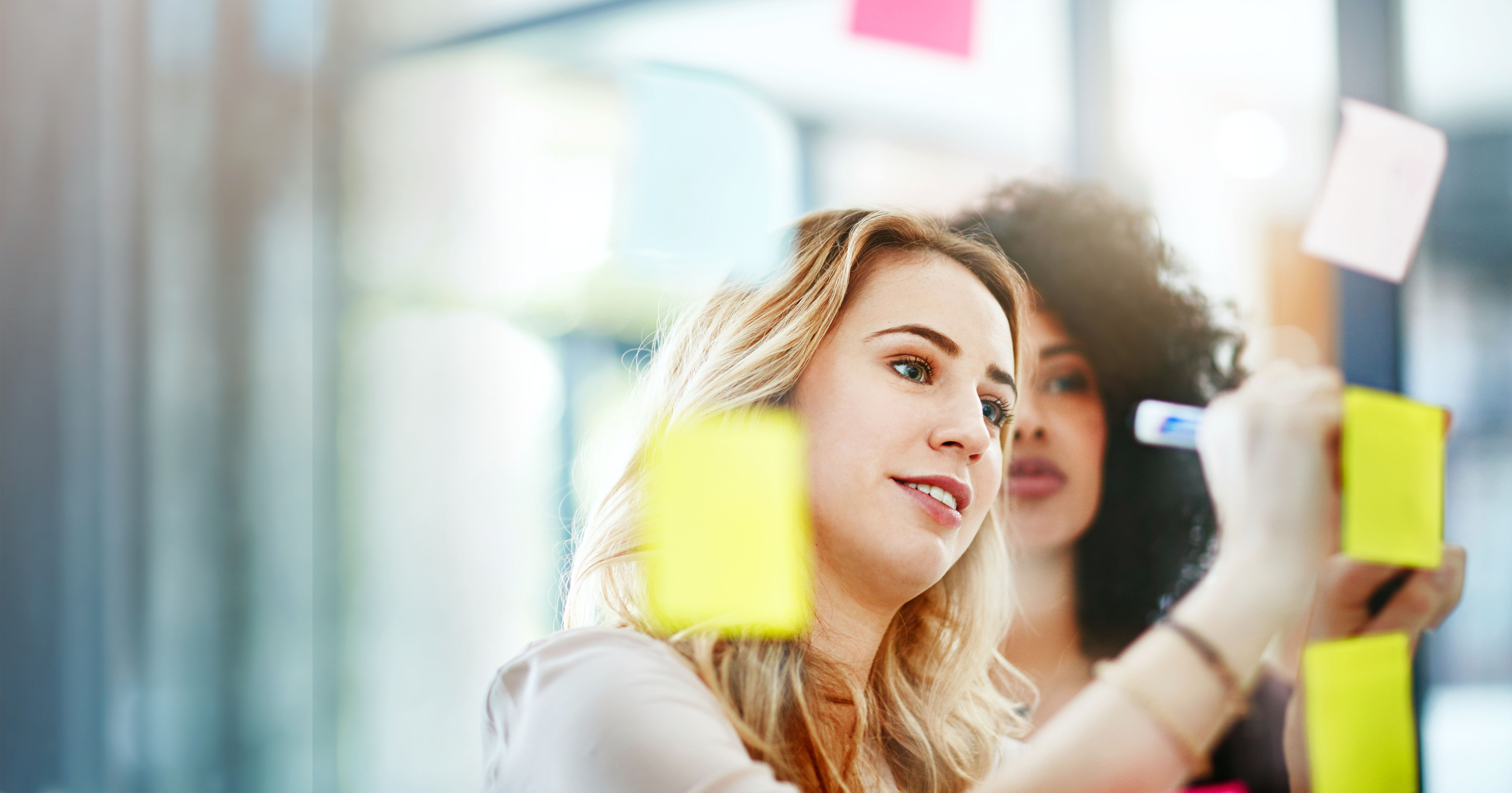 two-women-writing-on-glass-wall-with-colorful-sticky-notes.jpeg